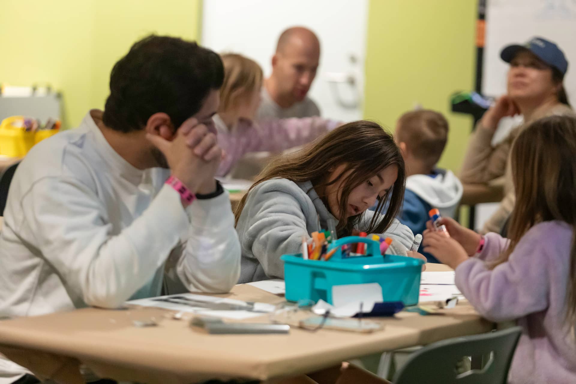 Drop-in Workshops For Families Family sitting at workshop craft table drawing with markers