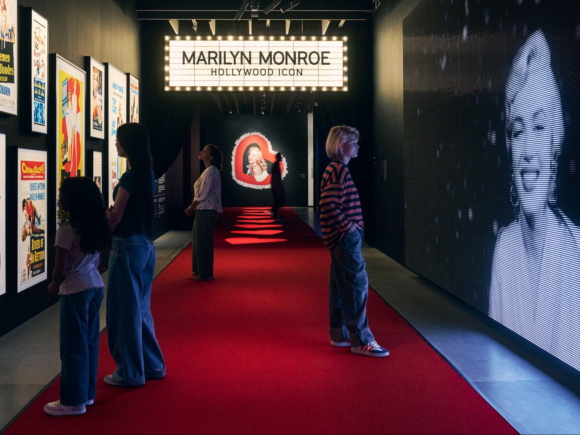 Visitors walk along a red carpet inside a museum exhibition dedicated to Marilyn Monroe. A large, illuminated marquee sign hangs overhead reading "MARILYN MONROE: HOLLYWOOD ICON," while vintage movie posters line the left wall and a massive digital screen on the right displays a black-and-white portrait of her smiling.