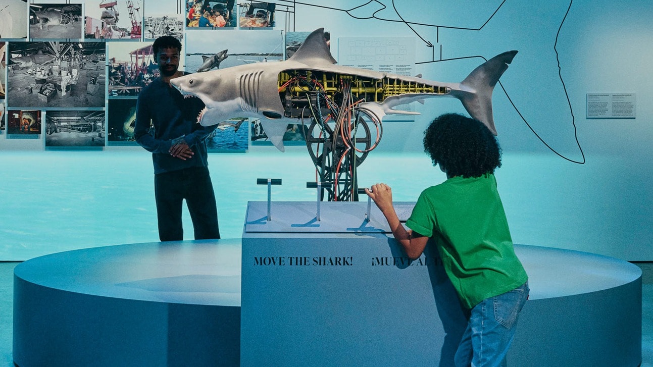 An image from an exhibit at the Academy Museum of Motion Pictures. The display is set in a dark room illuminated by blue lighting. The main wall features a large, schematic line drawing of a great white shark. Overlaid on the drawing is a collage of behind-the-scenes photographs related to the film Jaws and a three-dimensional, partially sectioned model of the mechanical shark, "Bruce," revealing its internal gears and mechanics. Three adult visitors stand on the left, looking at the display. In the foreground, a young boy in a green shirt and jeans is interacting with a control panel on a circular, multi-tiered pedestal in front of the model. The pedestal has text that reads, "MOVE THE SHARK! ¡MUEV..."