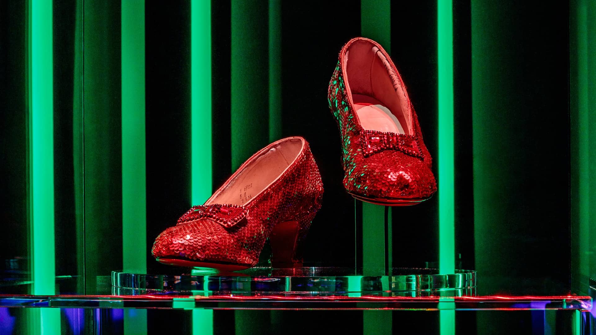 A pair of iconic ruby slippers, covered in red sequins with a matching bow on the toe, are displayed on clear pedestals. They are set against a dramatic backdrop of bright vertical green lights on a black background, reminiscent of the Emerald City from The Wizard of Oz.